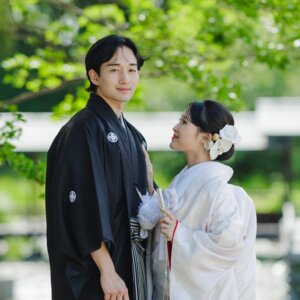 新緑の下で扇子を手に寄り添う黒紋付袴の新郎と白無垢の花嫁を縦位置でとらえた一枚。白鳥庭園で撮る和装前撮り