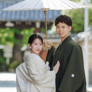 和傘の下で寄り添い、カメラにほほえむ白無垢の花嫁と紋付袴の新郎。名古屋で撮った和装の結婚写真
