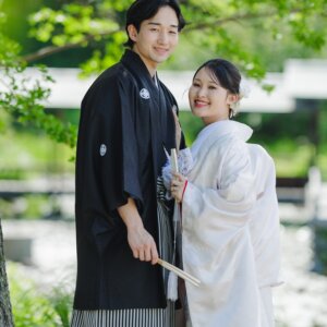 新緑の木々の下で笑い合う黒紋付袴の新郎と白い花の髪飾りの白無垢の花嫁。日本庭園で撮る和装ロケーションフォト