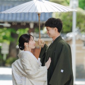 白い和傘を差す紋付袴の新郎と見つめ合う白無垢の花嫁。寺院の門を背景にした和装ロケーションフォト