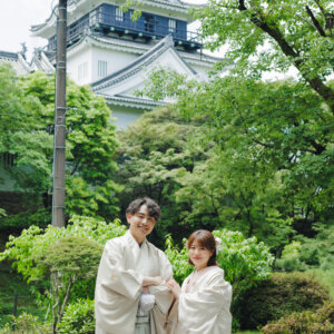 白い天守を背に立つ白い羽織袴の新郎と白い色打掛の花嫁を縦位置でとらえた一枚。岡崎城で撮る和装ロケーションフォト
