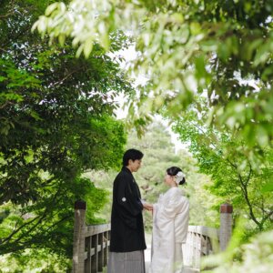 緑に囲まれた木の橋で向かい合う黒紋付袴の新郎と白無垢の花嫁を引きでとらえた一枚。白鳥庭園で撮る和装前撮り