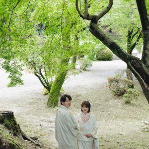 新緑の木々の下の小道を歩く白い羽織袴の新郎と白い色打掛の花嫁の後ろ姿。岡崎公園で撮る和装前撮り