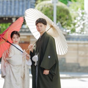 瓦屋根の門を背に、朱色と白の和傘を手にカメラを見つめる白無垢の花嫁と紋付袴の新郎。名古屋の和装ロケーション前撮り