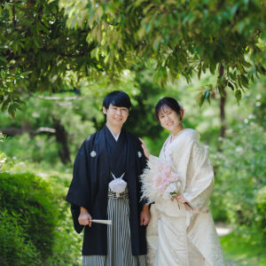 緑の木々の下でブーケを持って並ぶ黒紋付袴の新郎と白い色打掛の花嫁。日本庭園で撮る和装前撮り
