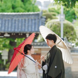 朱色の和傘の花嫁と白い和傘の新郎が、寺院の塀の前で向かい合う一枚。白無垢で撮る和装前撮り