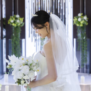 ロングベールとティアラを着けた花嫁が白いブーケを抱えてうつむく横顔。グリーンの装花がやわらかくボケたチャペルでの前撮り写真