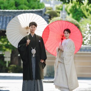 寺院の門の前で白と赤の和傘をそれぞれ差して並ぶ紋付袴の新郎と白無垢の花嫁。名古屋で撮る和装ロケーションフォト