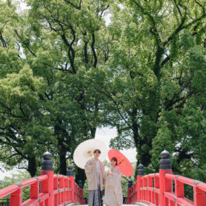 大木を背に、朱い橋の上で白と赤の和傘を差す白い羽織袴の新郎と白い色打掛の花嫁を引きでとらえた一枚。岡崎公園の朱い橋で撮る和装ロケーションフォト