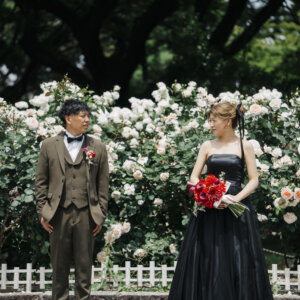 白い薔薇の生垣の前で離れて立つオリーブのスーツの新郎と赤いブーケを持つ黒いドレスの花嫁。庄内緑地の薔薇園で撮る洋装前撮り