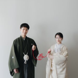 白い壁の前で赤い糸の両端を持ち、並んで笑う黒紋付袴の新郎と白無垢の花嫁。和装前撮りで人気の赤い糸のポーズ