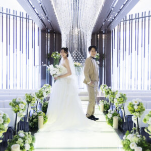 白いソファ席とグリーンの装花が並ぶ明るいチャペルで、背中合わせのように並ぶ新郎新婦。ロングトレーンのウェディングドレスが映えるチャペルフォト