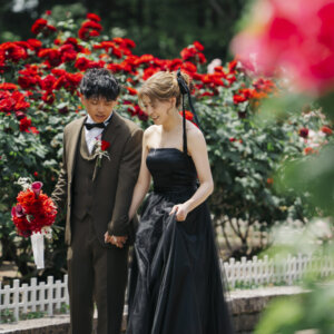 赤い薔薇を前ボケにして、手をつないで歩くオリーブのスーツの新郎と黒いドレスの花嫁。薔薇園で撮る洋装ロケーションフォト