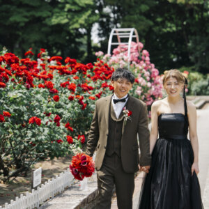 赤い薔薇の花壇の前で手をつないで歩くオリーブのスーツの新郎と黒いドレスの花嫁。庄内緑地の薔薇園で撮る洋装前撮り
