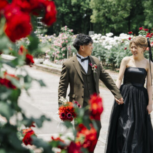 薔薇を前ボケにして手を取り合うオリーブのスーツの新郎と黒いドレスの花嫁。薔薇園で撮る洋装前撮り