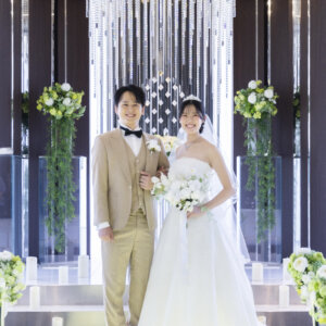 グリーンの装花が並ぶ祭壇の前で、腕を組んで微笑むベージュのタキシードの新郎とベール姿の花嫁。ラグナヴェール名古屋で撮る結婚写真