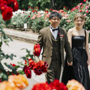 赤い薔薇の花壇の間の道を手をつないで歩くオリーブのスーツの新郎と黒いドレスの花嫁。庄内緑地の薔薇園で撮る洋装ロケーションフォト