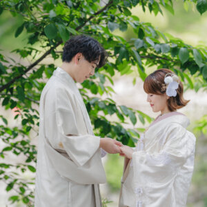 新緑の下で手を取り合う白い羽織袴の新郎と白い色打掛の花嫁を縦位置でとらえた一枚。公園で撮る和装ロケーションフォト