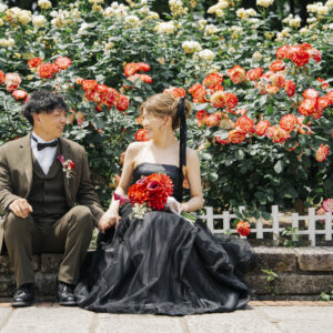 オレンジと黄色の薔薇の前の石段に座って見つめ合うオリーブのスーツの新郎と黒いドレスの花嫁。庄内緑地の薔薇園で撮る洋装前撮り