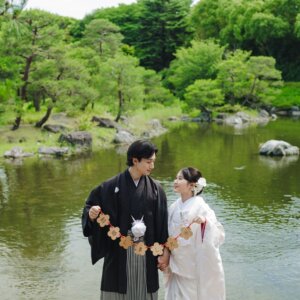 池のほとりで花のガーランドを持つ黒紋付袴の新郎と白無垢の花嫁。ガーランドを使った和装前撮りのアイテムカット