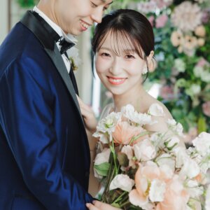 花の壁の前で頬を寄せ、ピンクのブーケを抱えてほほえむ新郎新婦のアップ。スタジオで撮る洋装の結婚写真