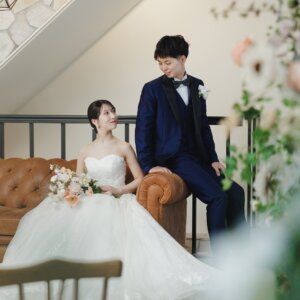 ソファの花嫁を見つめるネイビーのタキシードの新郎。草花越しに撮った名古屋のスタジオでの洋装フォトウェディング