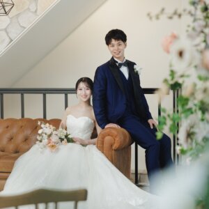 階段下のソファに座る花嫁と、ソファの端に腰を下ろす新郎。花で手前をぼかしたウェディングドレスのスタジオ前撮り