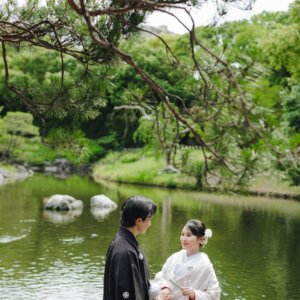 松の枝の下、池のほとりで向かい合う黒紋付袴の新郎と白無垢の花嫁。白鳥庭園で撮る和装ロケーションフォト