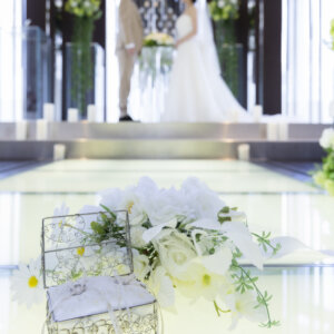 光る床のバージンロードに置いたガラスのリングピローと白い花のブーケ。奥の祭壇に立つ新郎新婦をぼかしたチャペルフォトの小物カット