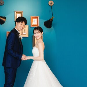ブルーの壁と金の額縁の前で手を取り合い、カメラに笑顔を向ける新郎新婦。カラフルな背景が映えるスタジオフォトウェディング
