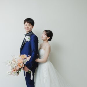 背中合わせに立つ新郎と、その背中からのぞき込む花嫁。白い背景で撮るウェディングドレスのスタジオ前撮りポーズ