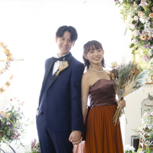 花のアーチとMR＆MRSのガーランドの前で手をつなぐ、ネイビーのタキシードの新郎とドライフラワーのブーケを持つカラードレスの花嫁の前撮り