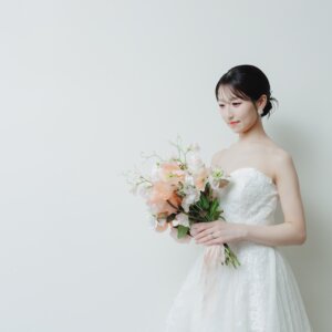 余白のある白い背景に、ブーケを持って目線を落とすウェディングドレスの花嫁。シンプルな構図のスタジオフォト