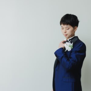 白い背景の前でブートニアに手を添え、うつむくネイビーのタキシードの新郎。スタジオで撮る新郎ソロのウェディングフォト