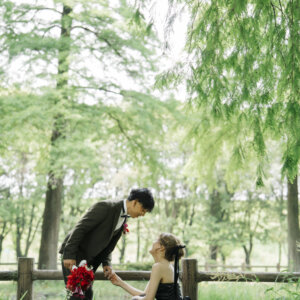 柵に座る黒いドレスの花嫁に赤いブーケを差し出すオリーブのスーツの新郎。庄内緑地で撮る洋装前撮りのプロポーズ風ポーズ