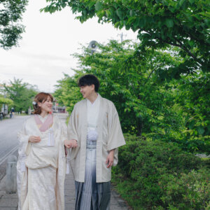 新緑の並木道を、白い羽織袴の新郎と白い色打掛の花嫁が並んで歩く一枚。岡崎公園で撮る和装ロケーションフォト