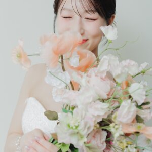 淡いピンクの花束に顔を寄せ、目を閉じてほほえむ花嫁のアップ。ブーケが主役のウェディングドレスのスタジオフォト