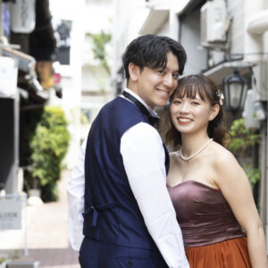 路地裏の街並みで寄り添ってカメラに笑いかける新郎新婦。パールのネックレスとテラコッタのカラードレスが映えるウェディングフォト