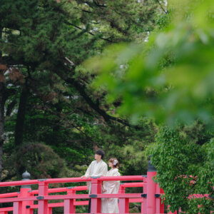 緑の木々に囲まれた朱い橋の上に立つ白い羽織袴の新郎と白無垢の花嫁を引きでとらえた一枚。岡崎公園の朱い橋で撮る和装前撮り
