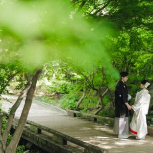 木漏れ日の木の橋で手を取り合う黒紋付袴の新郎と白無垢の花嫁を引きでとらえた一枚。日本庭園で撮る和装前撮り