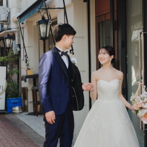 レンガ敷きの商店街で、ブーケを持つウェディングドレスの花嫁と手をつなぎ見つめ合う新郎。街並みで撮る洋装ロケーションフォト