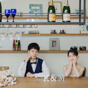 シャンパンの棚が並ぶカフェ風のセットで、テーブルに頬杖をつく新郎新婦。イニシャルの小物と一緒に撮る名古屋のスタジオ前撮り