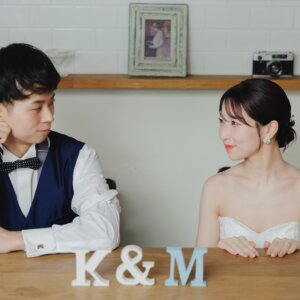 'K&M'のイニシャルオブジェを置いたテーブルで、頬杖をつき見つめ合う新郎新婦。カフェ風セットで撮るおしゃれなスタジオフォト