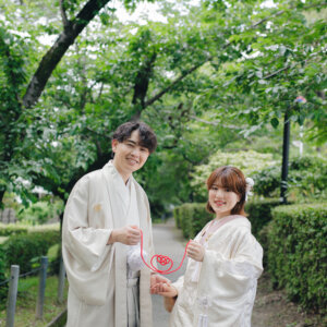 新緑の並木道で赤い糸を指にかけて見せる白い羽織袴の新郎と白い色打掛の花嫁。赤い糸を使った和装前撮りのアイテムカット