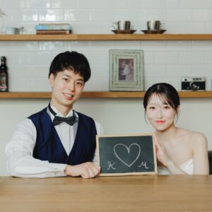 カフェ風のセットでハートを描いた黒板を手にほほえむベスト姿の新郎とウェディングドレスの花嫁。小物を使ったスタジオ前撮り