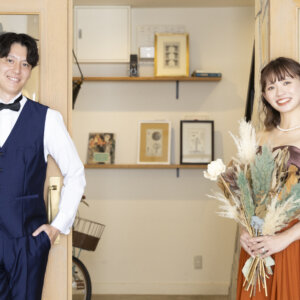木の扉と飾り棚のある部屋で、ネイビーのベストの新郎とドライフラワーのブーケを持つテラコッタのカラードレスの花嫁が笑顔を見せる名古屋のスタジオフォト