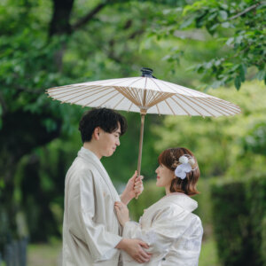 岡崎公園にて、白い和傘を差して寄り添う白い羽織袴の新郎と白い色打掛の花嫁を縦位置でとらえた一枚。新緑の公園で撮る和装前撮り