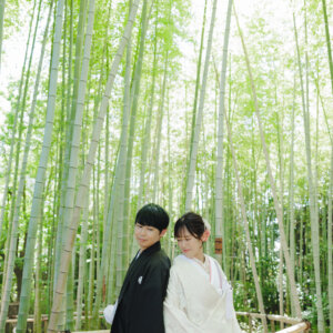竹林の柵のそばに並んで立つ黒紋付袴の新郎と白い色打掛の花嫁を縦位置でとらえた一枚。竹林で撮る和装ロケーションフォト