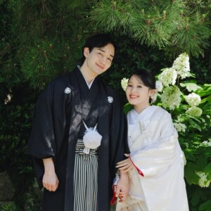 松と白い花を背に腕を組んで並ぶ黒紋付袴の新郎と白無垢の花嫁。白鳥庭園で撮る和装前撮りの正面カット