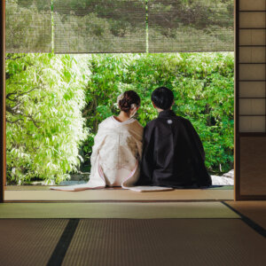簾の下がる和室から庭を眺める黒紋付袴の新郎と色打掛の花嫁の後ろ姿。白鳥庭園の日本家屋で撮る和装前撮り